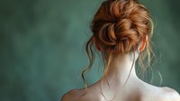 Elegant messy bun hairstyle with loose ginger strands on pale skin against green background, view from behind showing nape and bare shoulders.
