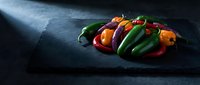 Colorful assortment of fresh hot chili peppers on dark slate board, dramatic moody lighting, rich green, red, orange and purple tones.