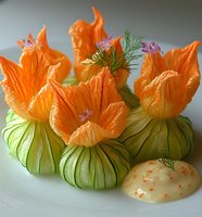 Creative appetizer made with carved cucumber and carrot flowers garnished with fresh dill and pink blossoms, served with citrus sauce on white plate.