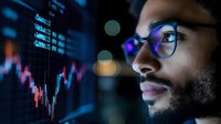 Young African American businessman in glasses analyzing financial market data on digital screen with stock charts and trading information in dark office environment.