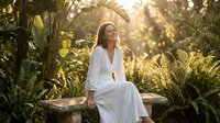Young woman in white dress sitting peacefully in tropical garden during golden hour sunset with lush green plants and warm natural lighting.