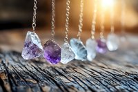 Raw crystal gemstone pendants hanging on silver chains with purple amethyst and clear quartz stones on rustic wooden surface with warm sunlight.