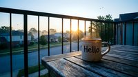 Steaming coffee mug with Hello text on wooden balcony railing at golden sunrise over suburban neighborhood in morning light.