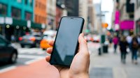 Hand holding smartphone with blank screen on busy city street with blurred urban background and colorful buildings for mobile app mockup and digital communication.