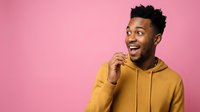 Young man with dark skin tone and black hair wearing a mustard yellow hoodie, smiling with hand near face against pink background for lifestyle and portrait use.