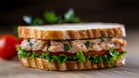 Fresh tuna salad sandwich with crisp lettuce on toasted bread, garnished with cherry tomatoes and herbs against rustic wooden background. Perfect for menu photography.