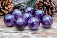 Purple amethyst crystal spheres arranged on rustic wooden surface with pine cones and evergreen branches, showcasing natural healing stones and spiritual elements.