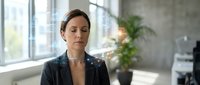 Professional businesswoman with closed eyes practicing mindfulness meditation in modern office space with natural light and plants for stress relief.
