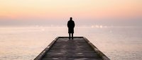 Silhouetted male figure standing alone on wooden pier at sunrise, gazing toward calm ocean horizon during golden hour, conveying solitude, contemplation, and peaceful morning reflection.
