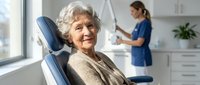 Elderly Caucasian woman smiling in dental chair while female dentist prepares equipment in modern clinic. Senior patient receiving professional oral care.