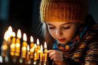 Young woman in orange knit beanie and scarf gazing thoughtfully at glowing Hanukkah menorah candles in dark room, celebrating Jewish holiday tradition.