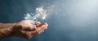 Hands holding glowing light bulb with sparkling particles dispersing into air against blue background, representing innovation, ideas, and energy solutions.