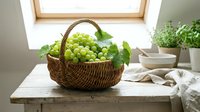 Green grapes in woven basket on wooden table by window with natural light, suitable for food, harvest, and kitchen styling projects.