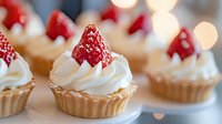 Fresh strawberry tartlets with whipped cream on golden pastry shells, arranged on white serving plate with soft bokeh background.