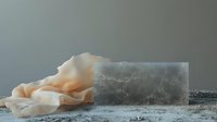 Minimalist composition with translucent selenite crystal block and delicate cream-colored fabric on textured surface against moody gray background, creating ethereal atmosphere.
