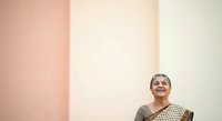 Senior woman with gray hair and glasses wearing traditional patterned clothing, looking upward against minimalist pastel wall backdrop with pink and cream sections.