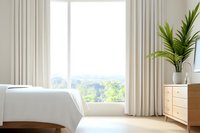 Bright minimalist bedroom interior with white bedding, cream curtains, wooden furniture and potted palm plant against panoramic window with forest view.