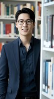 Young East Asian male professional wearing eyeglasses and navy blazer over dark shirt, smiling in library setting with bookshelves. Suitable for business, education, and corporate communications.