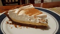 Slice of layered cream pie with chocolate cookie crust, caramel filling, and whipped cream topping dusted with cocoa powder, served on white plate with blue rim.