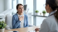 Middle-aged Caucasian woman patient consulting with female doctor in modern medical office, healthcare appointment discussion about symptoms.