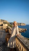 Elegant mature Caucasian woman in flowing white dress standing on Mediterranean coastal terrace with blue sea and cliffs backdrop during golden hour.