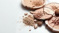Broken brown makeup powder compacts with scattered cosmetic dust and fragments on white background for beauty product photography.