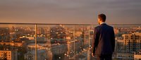 Young businessman in dark suit standing on modern office balcony overlooking urban cityscape at golden hour sunset, contemplating future success and opportunities.