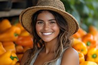 Young Hispanic woman with straw hat smiling at farmers market with fresh orange bell peppers in background, representing healthy lifestyle and local produce.