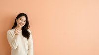 Young Asian woman with long dark hair wearing a cream-colored sweater, smiling and touching her face against a warm peach background. Suitable for lifestyle, beauty, and wellness marketing materials.