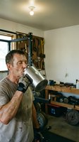Middle-aged male worker drinking from metal thermos in workshop garage. Caucasian man wearing work gloves and apron taking break surrounded by tools and equipment on shelves.