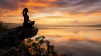 Woman in silhouette sitting on rocky cliff overlooking calm lake at golden sunrise, meditating in peaceful natural landscape with misty water reflection.