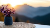 Purple wildflowers in blue knitted vase on wooden table against mountain sunset backdrop, creating serene outdoor atmosphere with warm golden light.