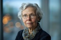 Senior Caucasian woman with silver hair and glasses wearing navy sweater and patterned scarf, gentle smile against soft bokeh background, professional portrait lighting.