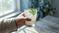 Hand holding a refreshing glass of lemonade with fresh mint leaves and ice cubes on a sunny windowsill, perfect for beverage, summer drink, and lifestyle content.