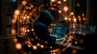 Hooded hacker in dark room working on laptop with glowing code and digital particles. Cybersecurity threat concept for technology and data protection.