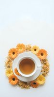 Overhead view of white cup filled with herbal tea surrounded by fresh and dried yellow and orange gerbera daisies on neutral background, ideal for wellness and beverage marketing.