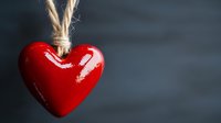 Bright red glossy heart ornament hanging on rustic twine string against dark moody background, symbolizing love, romance and Valentine's day celebration.