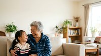 Elderly woman with gray hair and Asian ethnicity embracing young granddaughter on beige sofa in bright living room with plants and natural light, depicting family bonding and intergenerational love.