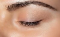 Close-up of a closed eye with natural makeup, showing defined eyebrow, subtle eyeshadow, and long eyelashes on smooth skin.