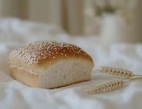 Fresh baked white bread loaf with sesame seeds on top and golden wheat ears on white linen cloth background, soft natural lighting creates cozy atmosphere.