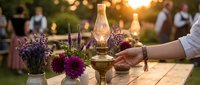 Elegant outdoor dinner party setup with vintage oil lamp, purple flowers and warm golden hour lighting creating romantic dining atmosphere.