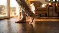 Barefoot person walking on warm wooden floor at home, cozy living room with natural light in background.