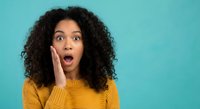 Young African American woman with curly hair showing shocked surprised expression touching face in yellow sweater on turquoise background for advertising.