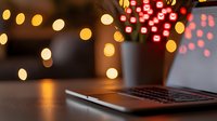 Laptop keyboard in cozy evening atmosphere with blurred Christmas lights and decorative red flowers creating warm bokeh effect in background.