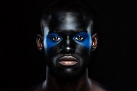 Young African American male model with bright blue face paint across eyes on black background, dramatic studio portrait with artistic makeup for fashion and beauty concepts.