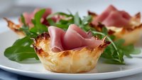 Crispy phyllo dough cups filled with prosciutto ham and fresh arugula, served as elegant appetizers on white plate. Perfect for gourmet catering events.