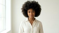 Young African American businesswoman with curly afro hair wearing white shirt in modern office space looking confident and professional.