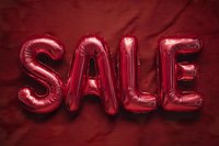Shiny red foil balloons spelling SALE against deep burgundy fabric background, creating bold contrast for retail promotion.