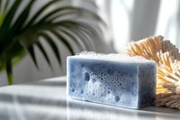 Blue bar soap with foam bubbles on marble surface, natural brush and plant in background, soft sunlight, skincare and hygiene concept.
