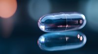 Shiny metallic capsule pill with reflection on mirror surface against blurred bokeh background, dramatic medical and pharmaceutical concept macro photography.